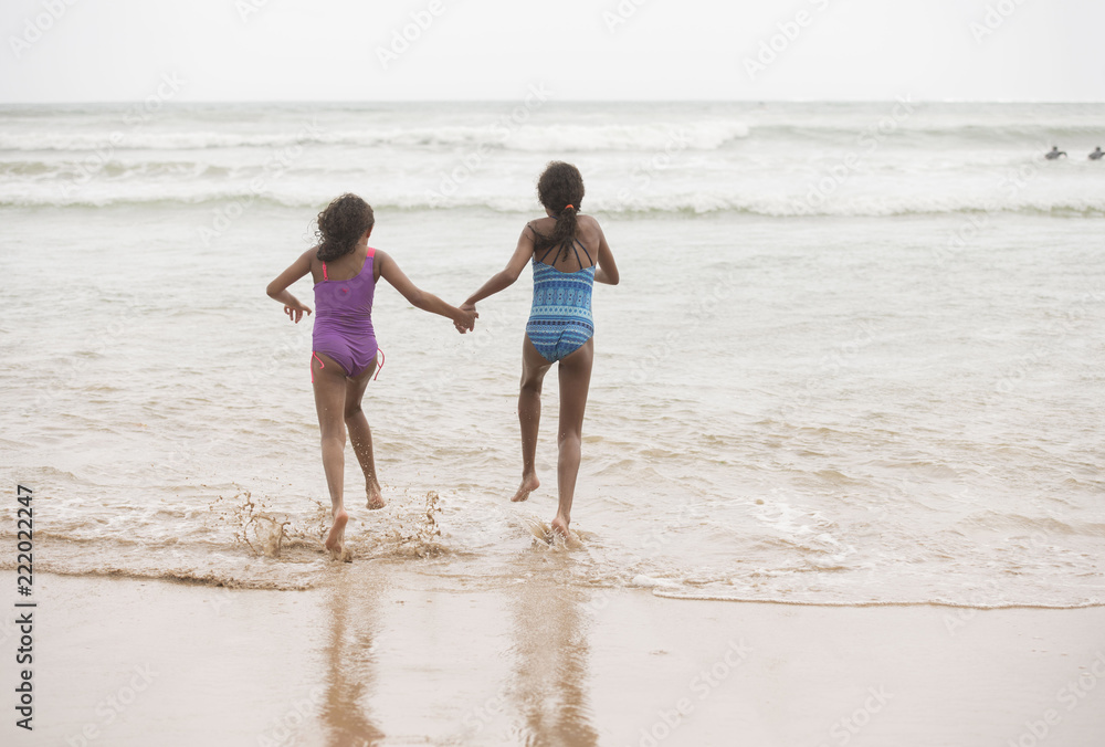 Rear view of two mixed-race sisters running into the water playing and laughing having fun on the beach on a bright tropical summer holiday