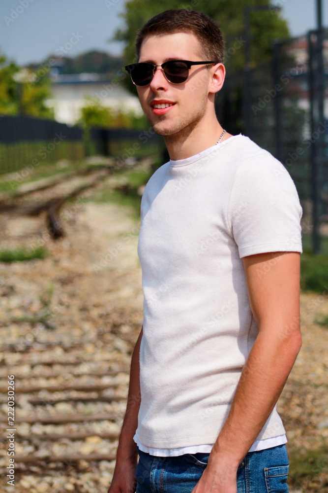Handsome young guy with sunglasses posing and enjoying a nice sunny day. Urban city environment. Old railway in background. Male model photo shoot, urban fashion, lifestyle. White T-shirt and jeans. Handsome young guy with sunglasses posing and enjoying a nice sunny day. Urban city environment. Old railway in background. Male model photo shoot, urban fashion, lifestyle. White T-shirt and jeans.