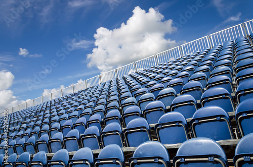 Seats in stadium, sky with clouds in background. Open air theater with natural daylight. Plastic blue seats rows as repeating pattern.