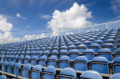 Seats in stadium, sky with clouds in background. Open air theater with natural daylight. Plastic blue seats rows as repeating pattern.