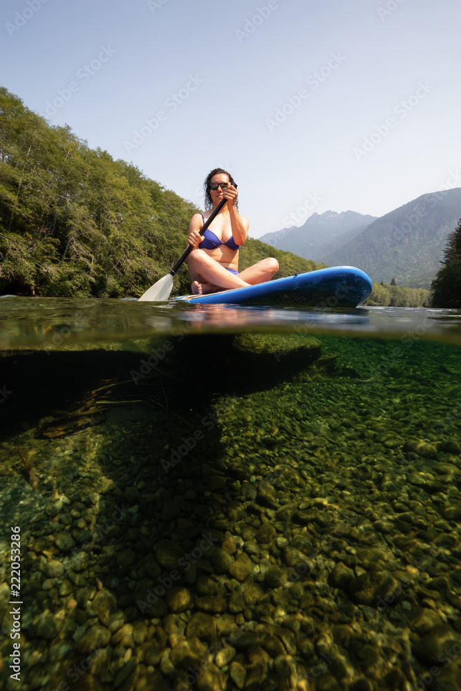 Over and Under picture of a woman paddle boarding in a river during a vibrant sunny summer day. Taken near Tofino and Ucluelet, Vancouver Island, BC, Canada.