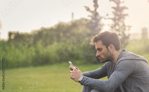 Happy young man browsing social media on his phone, sitting outside in a park, typing a message to a friend