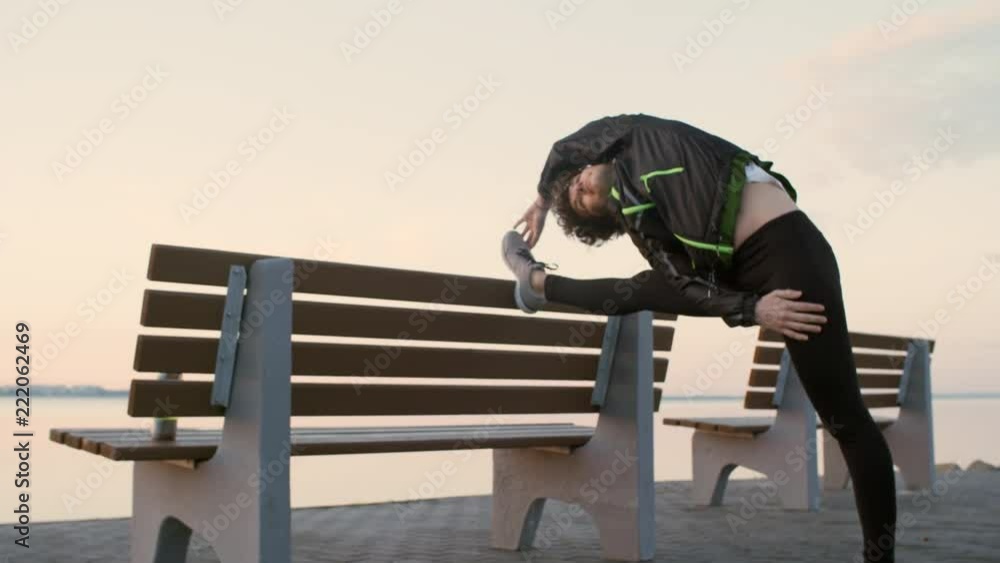 Zoom in of young athletic woman in sportswear stretching her legs on bench while warming up before morning workout on lakeside promenade Zoom in of young athletic woman in sportswear stretching her legs on bench while warming up before morning workout on lakeside promenade