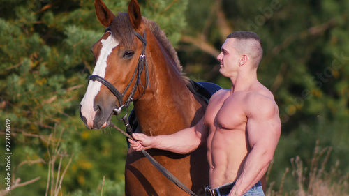 Athletic man with bare torso in denim pants standing next to the horse, holding the horse by the bridle