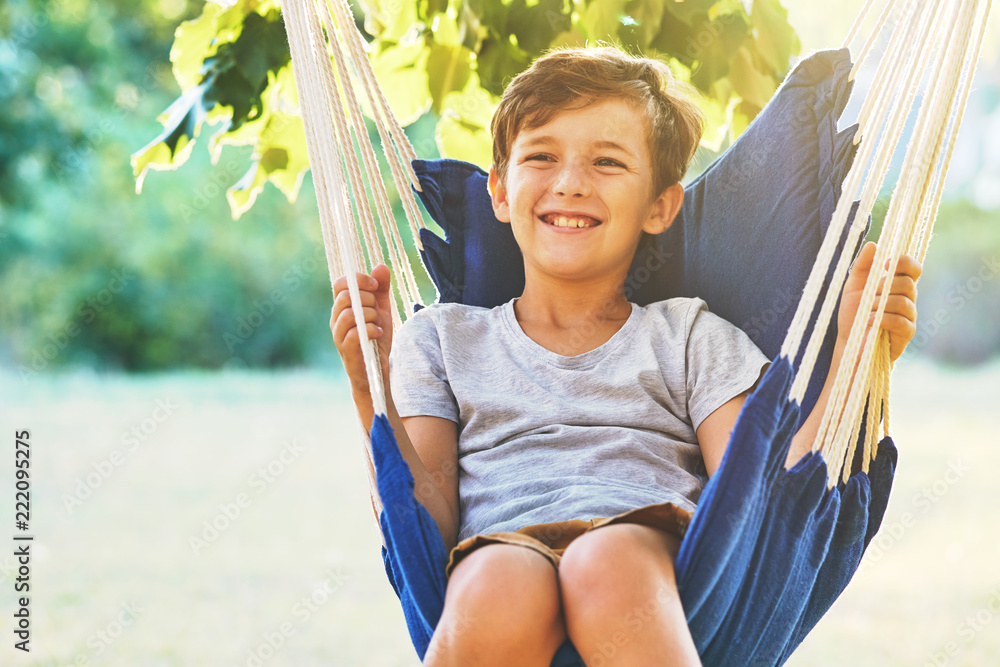 Autumn greeting card with place for your text. A happy boy sits in a comfortable blue hammock that hangs on a tree in the garden on bright background of green foliage, sun rays and autumn trees. Relax