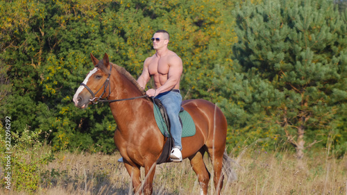 Brutal strong young man rides a horse on the field in the summer