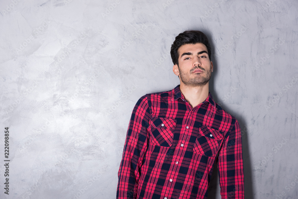 A serious handsome young man in a red checkered shirt standing in front of a grey wall in a studio. A serious handsome young man in a red checkered shirt standing in front of a grey wall in a studio.