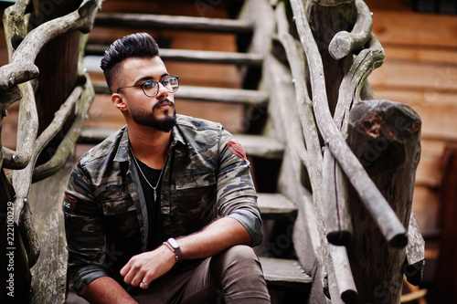 Awesome beautiful tall ararbian beard macho man in glasses and military jacket posed outdoor against wooden stairs. © AS Photo Family