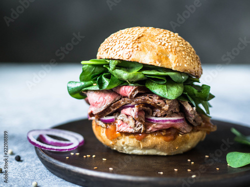 sandwich with steak slices, sauce of salsa, spinach and red onion. Burger from a bun with sesame and steak on a wooden plate on a gray background

