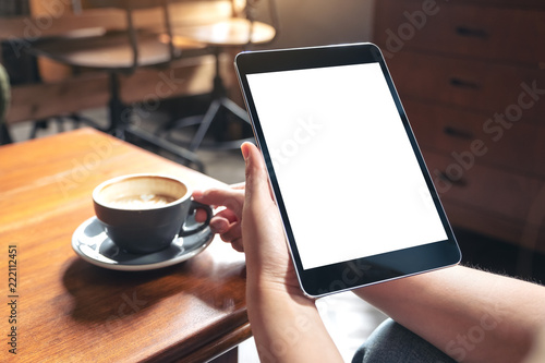 Mockup image of woman's hands holding black tablet pc with blank white screen while drinking coffee on wooden table in cafe