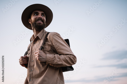 Portrait of young bearded man with optimistic smile admire beautiful nature and sunset. Handsome traveller wearing hipster hat and leather backpack walking alone. Time to travel concept.