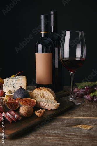 close-up view of bottles and glass of red wine, fruits and delicious cheese on wooden table