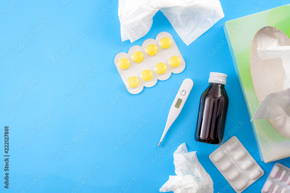 Sick day and medical leave concept with flat lay of cough syrup, thermometer, sore throat lozenges, bos of paper tissues, pills and medicines isolated on blue background with copy space