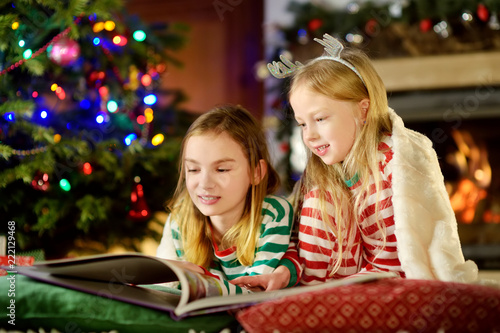 Happy little sisters reading a story book together by a fireplace in a cozy dark living room on Christmas eve.