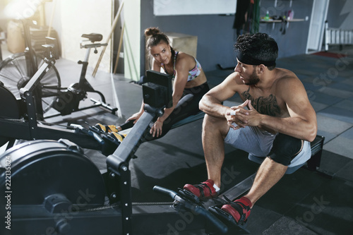 Gym. Man And Woman Training On Rowing Machine At Crossfit Club 