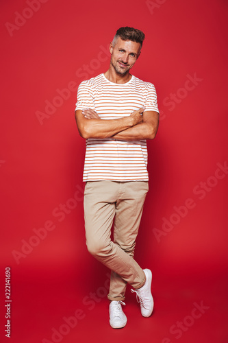 Full length photo of attractive man in striped t-shirt smiling and standing with arms crossed, isolated over red background