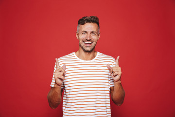 Positive man in striped t-shirt gesturing index fingers upward at copyspace, isolated over red background