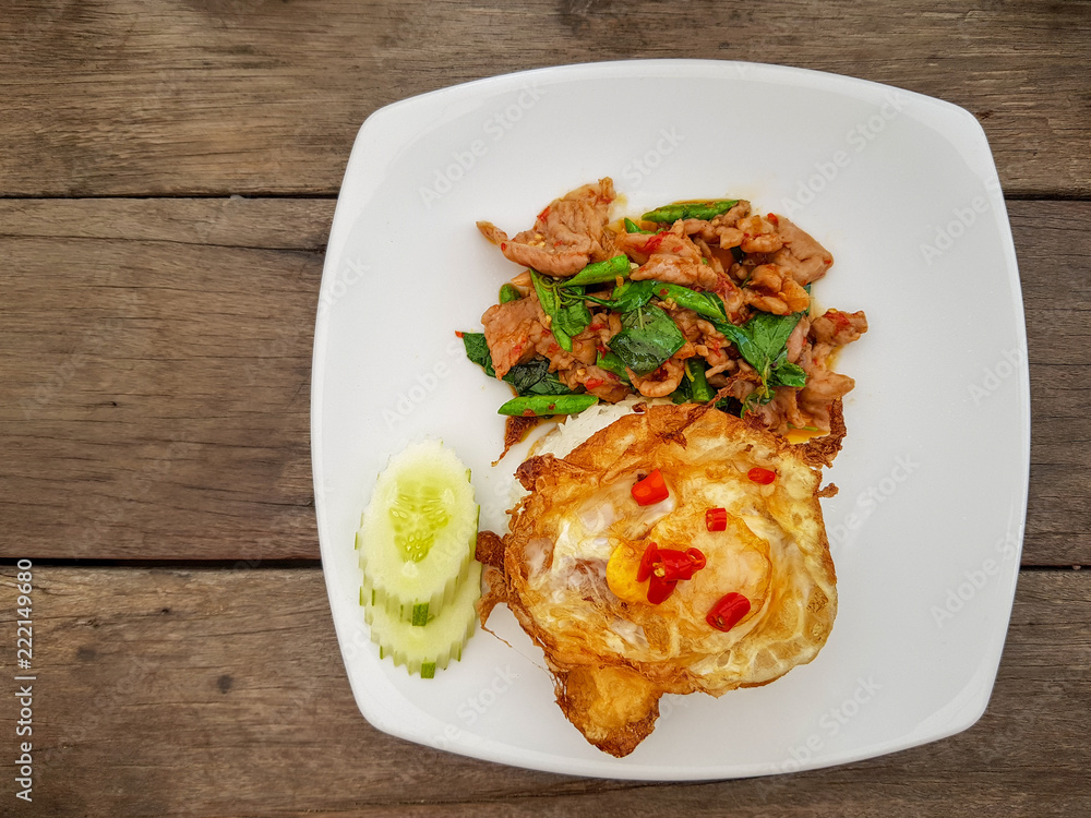 Hot and spicy fried fermented pork with , basils and green long beans served with white rice, crispy fried egg , sliced cucumber and red chillies on white dish over wooden table. Hot and spicy fried fermented pork with , basils and green long beans served with white rice, crispy fried egg , sliced cucumber and red chillies on white dish over wooden table.