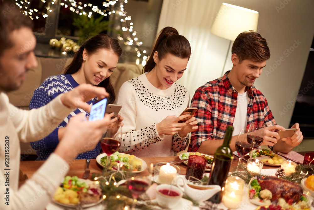 holidays and celebration concept - happy friends with smartphones having christmas dinner at home