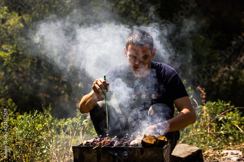 a young male guy preparing a shish kebab out of meat and chicken on nature in the mountains a lot of smoke
