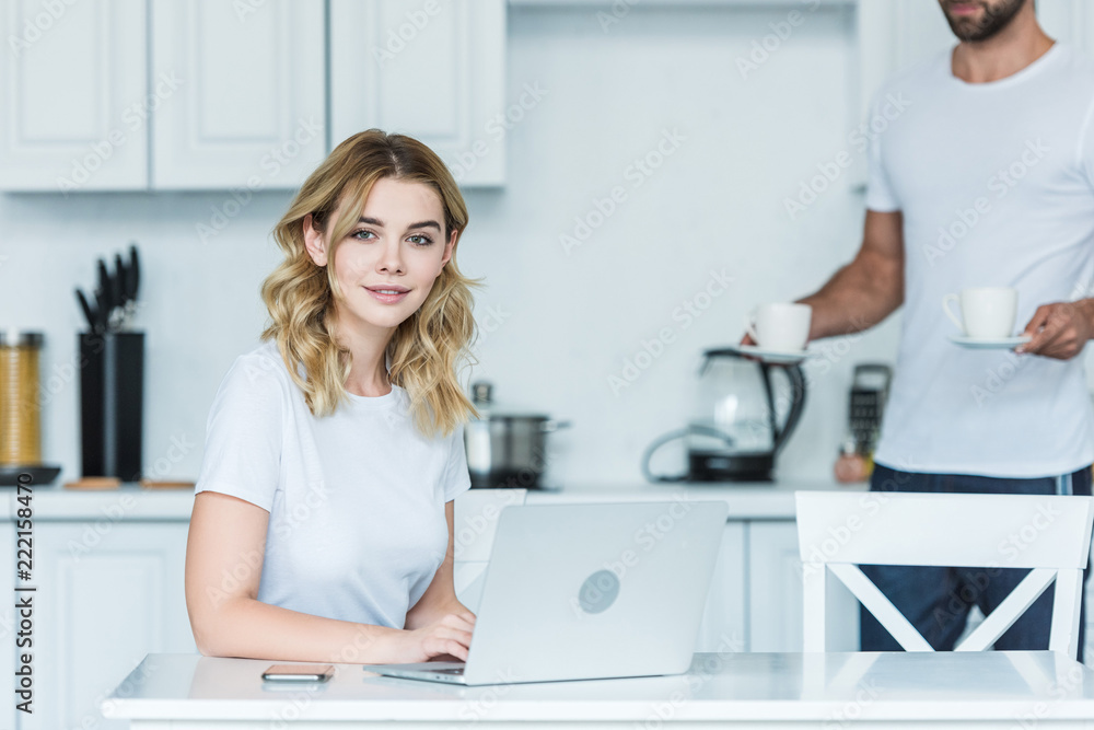 young woman using laptop and smiling at camera while boyfriend holding cups of coffee behind young woman using laptop and smiling at camera while boyfriend holding cups of coffee behind