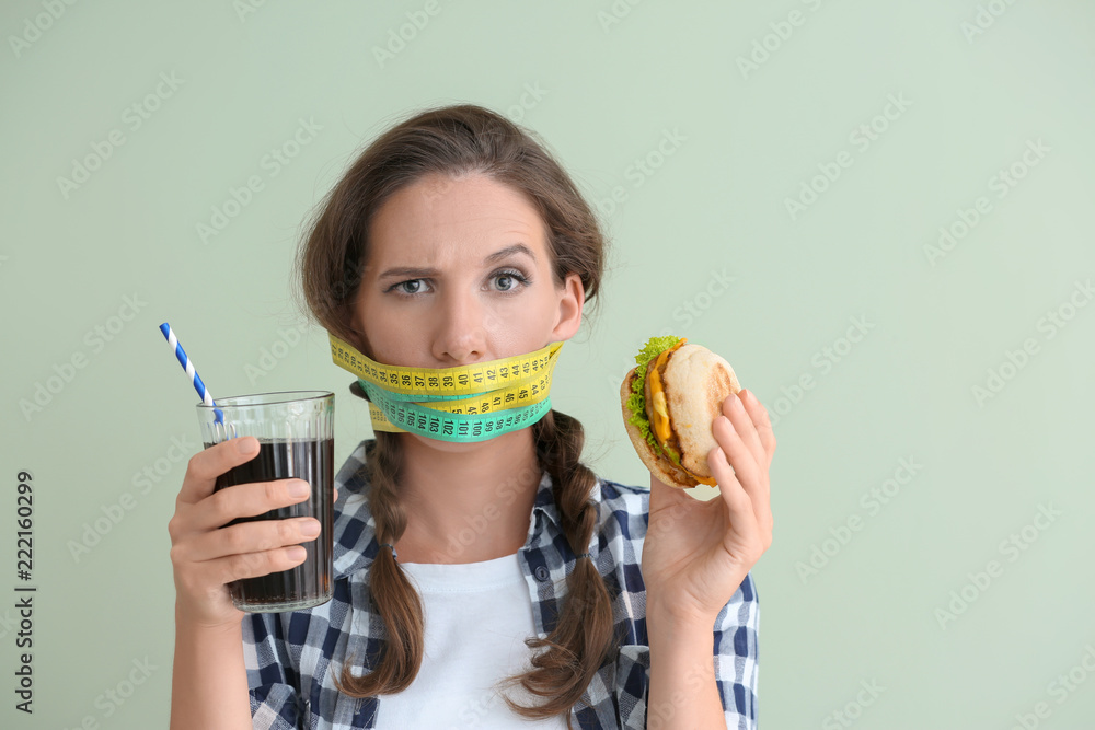 Emotional woman with measuring tapes around her mouth and unhealthy food on color background. Diet concept - obrazy, fototapety, plakaty 