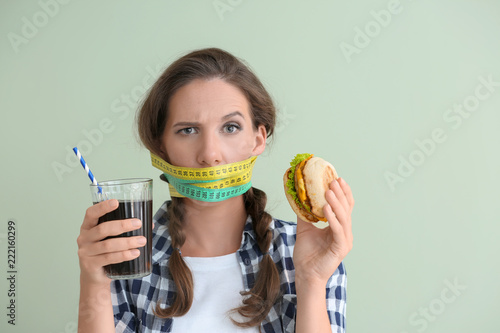 Emotional woman with measuring tapes around her mouth and unhealthy food on color background. Diet concept