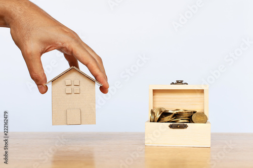 Hand holding house model and money coins in wood box on wooden table. Concept for property, mortgage, saving and real estate investment