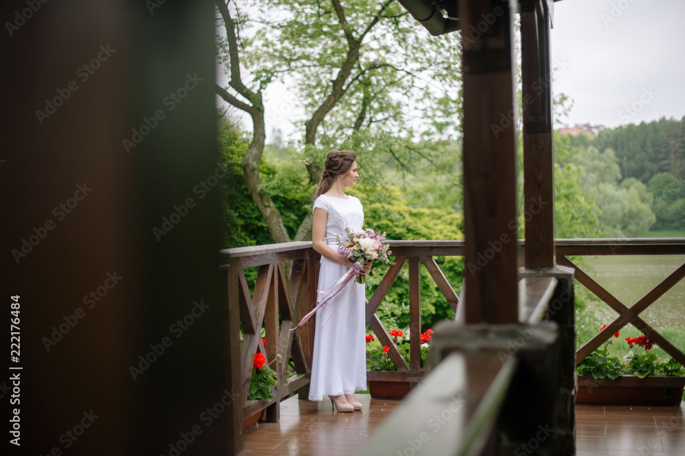 Young bride in wedding dress standing on the terrace of the house. Tender woman on the porch on the wedding day. Preparing for the ceremony.