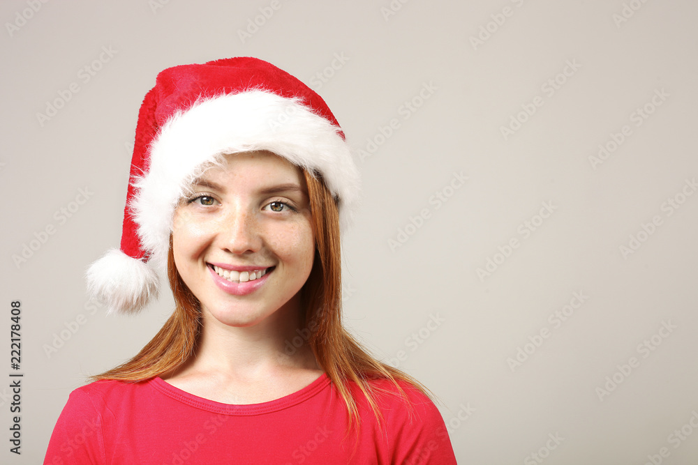 Close up portrait of beautiful redheaded young woman wearing Santa Claus hat and red t-shirt with satisfied facial expression. Cute female celebrating New Year. Copy space, isolated on gray background Close up portrait of beautiful redheaded young woman wearing Santa Claus hat and red t-shirt with satisfied facial expression. Cute female celebrating New Year. Copy space, isolated on gray background