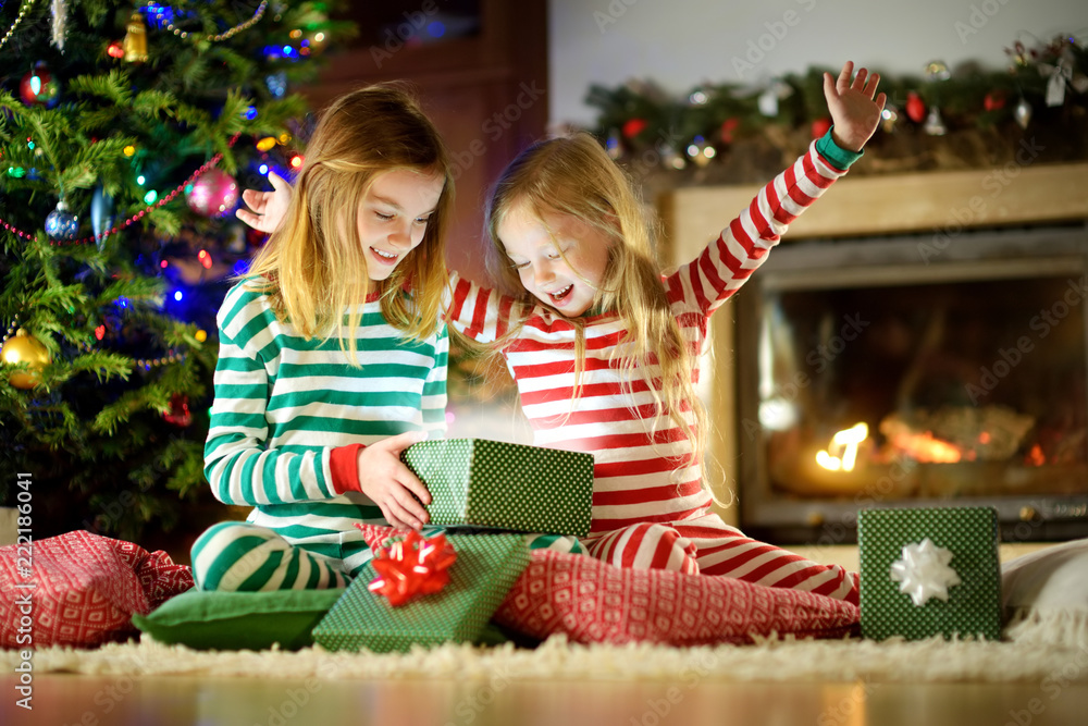 Happy little sisters wearing Christmas pajamas opening gift boxes by a fireplace in a cozy dark living room on Christmas eve. Happy little sisters wearing Christmas pajamas opening gift boxes by a fireplace in a cozy dark living room on Christmas eve.