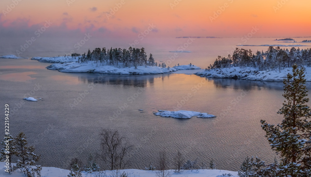 Fototapeta premium A colorful sunset on the Ladoga lake in the frost. Winter day. Open water hovers from frost. Islands in the snow.