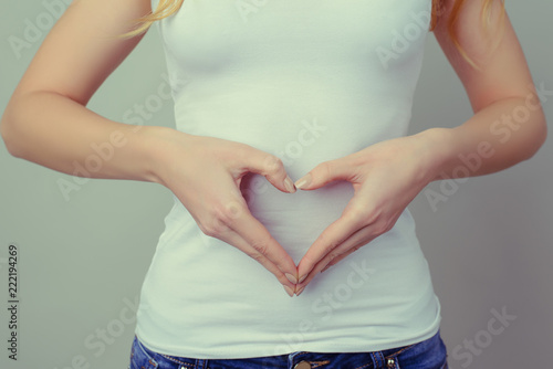 Concept of first weeks pregnancy. Woman's hands forming heart on her tummy. Concept of healthy eating and lifestyle. Woman in white t-shirt and jeans isolated against grey background photo