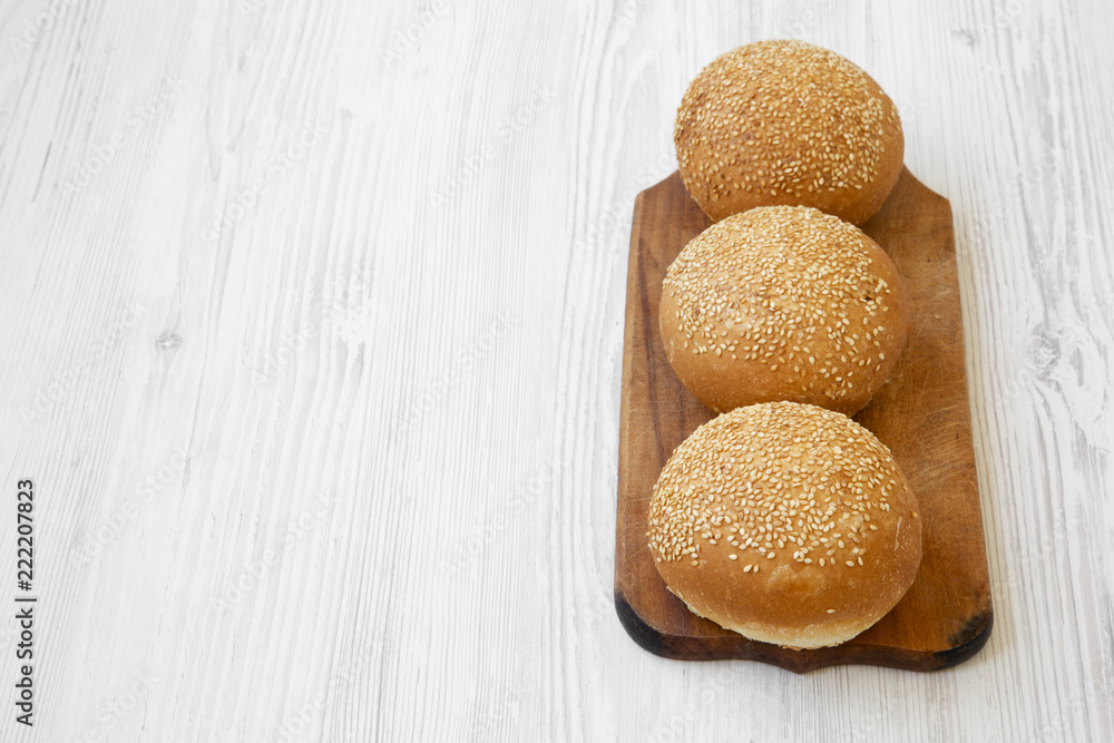 Fresh burger buns with sesame seeds on wooden board on white wooden table, high angle view. Fast food. Space for text. Fresh burger buns with sesame seeds on wooden board on white wooden table, high angle view. Fast food. Space for text.