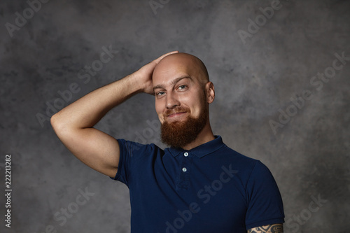 Picture of handsome attractive young bearded man wearing stylish polo shirt looking at camera with flirty expression, scratching his shaved bald head, feeling shy while talking to beautiful woman