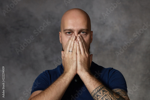 Portrait of attractivebald man with tattoo covering mouth and nose with both hands, holding breath because of bad smell. Tired exhausted frustrated male posing in stduio, keeping hands on his face photo
