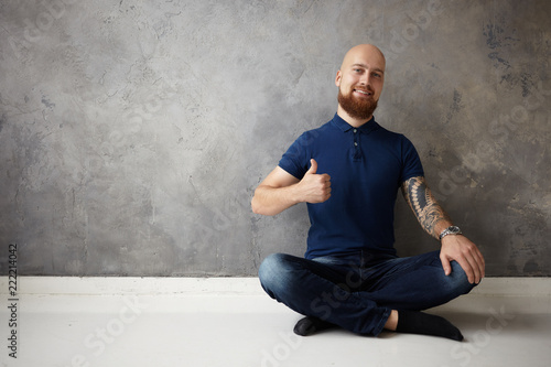 Picture of handsome positive young bald bearded guy with tattoo on his muscular arm smiling broadly at camera and making thumbs up gesture, having great day, being in good mood, showing approval