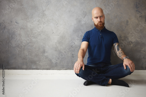 Human facial expressions and body language. Emotional grumpy young unshaven bearded male with shaved head sitting on floor keeping legs crossed, being angry while can't relax, trying to meditate