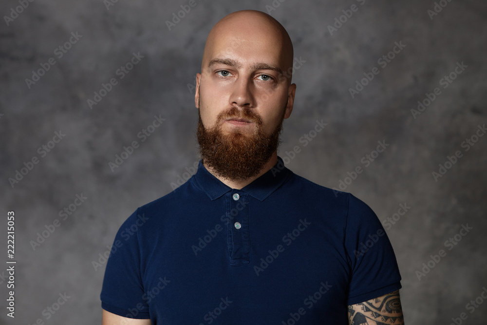 Horizontla shot of gloomy young bald male with bushy beard being dissatisfied with unfair results of competition, pursuing lips, trying to hide his anger, staring at camera with sullen annoyed look