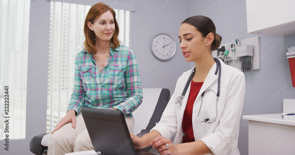 Portait of young latina doctor using laptop computer while talking with senior patient inside clinic. Close up of middle aged patient discussing with young female doctor health issues Portait of young latina doctor using laptop computer while talking with senior patient inside clinic. Close up of middle aged patient discussing with young female doctor health issues