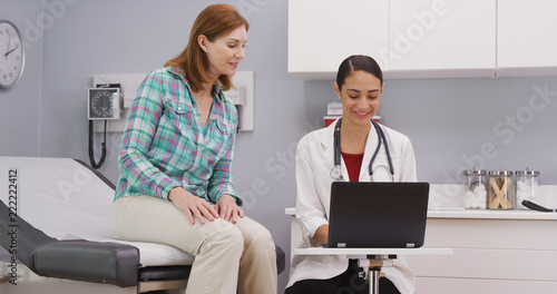 Lovely young medical doctor attending to mid aged patient using laptop computer. Senior caucasian patient consulting with hispanic doctor about health condition