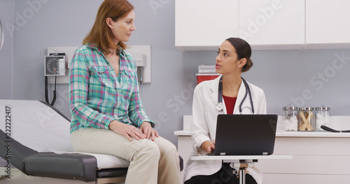 Healthy middle aged patient consulting with latina doctor about health test results. Young beautiful hispanic doctor using notebook computer to review patients health history