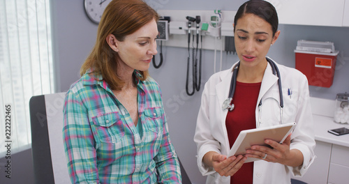 Charming female senior patient consulting with young latina doctor about health condition. Portrait of young hispanic doctor using portable tablet to look at test results of mid aged patient