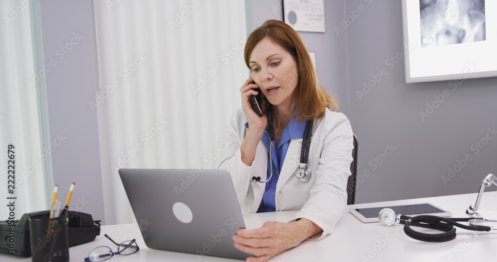 Attractive mid aged doctor using latop computer while talking on smartphone with colleague. Charming senior physician sitting in her office looking at notebook computer and chatting on cell phone Attractive mid aged doctor using latop computer while talking on smartphone with colleague. Charming senior physician sitting in her office looking at notebook computer and chatting on cell phone