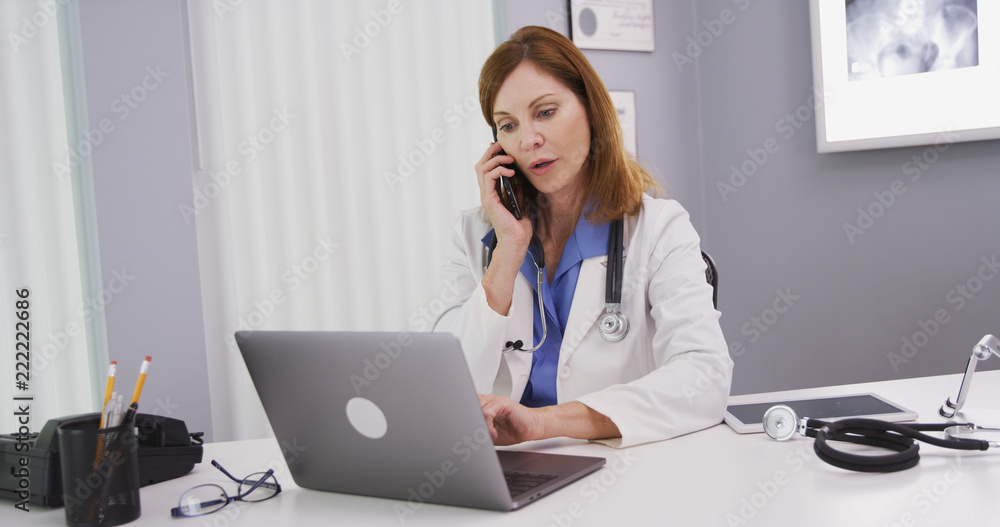 Attractive mid aged doctor using latop computer while talking on smartphone with colleague. Charming senior physician sitting in her office looking at notebook computer and chatting on cell phone Attractive mid aged doctor using latop computer while talking on smartphone with colleague. Charming senior physician sitting in her office looking at notebook computer and chatting on cell phone