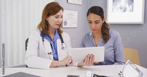 Senior medical physician using high-tech tablet to share lab results with young patient. Young patient consulting with mid aged doctor her health conditions looking at tablet computer