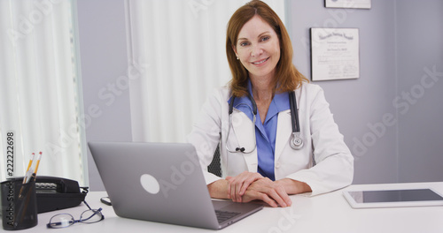 Portrait of lovely female doctor sitting at office desk smiling at camera. Close up of professional medical physician looking at camera