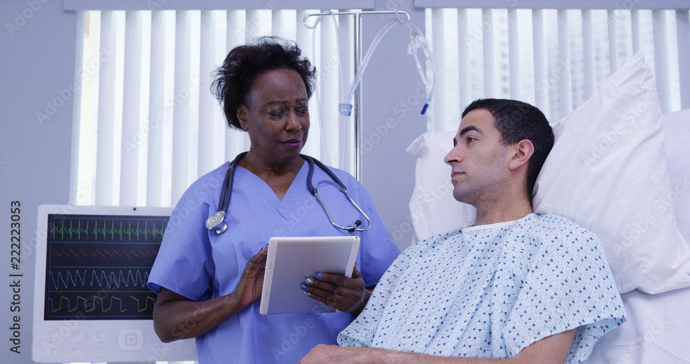 Obraz premium Mid aged African nurse checking in on young latino patient with electronic notebook tablet. Charming male patient lying down hospital bed with senior nurse attending using portable electronic pad