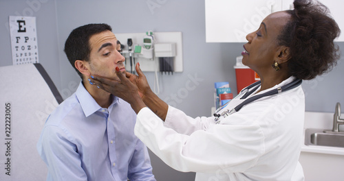 Professional black MD checking young patients neck for swollen thyroid and lymph nodes. Young latino male patient having routine health checkup