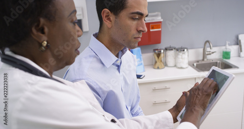 Close-up of mid aged blact doctor using portable tablet to show patient his xray results. Attractive latino patient looking at x-ray result of his neck and spine on electronic notebook tablet
